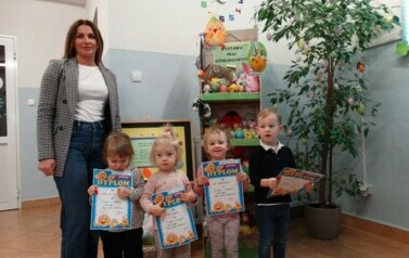 Na pierwszym planie stoi Pani Dyrektor z czw&oacute;rką dzieci. W ręce trzymają dyplomy. W tle regał z kolorowymi pisankami. 