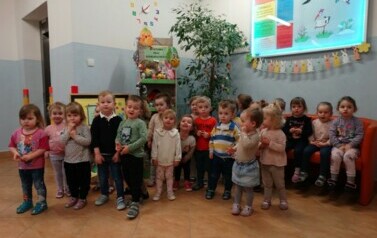 Na pierwszym planie stoi grupa dzieci. W tle regał z kolorowymi pisankami. 