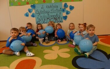 Sala zabaw-grupa dzieci siedzi na dywanie. W ręce trzymają niebieskie balony. W tle jasna ściana a na niej niebieskie serduszka. 