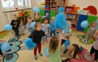 Sala zabaw-grupa dzieci stoi na dywanie. W ręce trzymają niebieskie balony i pompony.