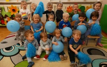 Sala zabaw-grupa dzieci siedzi na dywanie. W ręce trzymają niebieskie balony. 