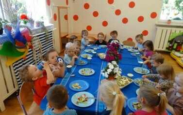Grupa dzieci siedzi przy stoliku, na kt&oacute;rym stoją kolorowe kwiaty i salaterki z sałatką.