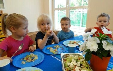 Czw&oacute;rka dzieci siedzi przy stoliku. Dziewczynki trzymają w ręce łyżki, kt&oacute;rymi dotykają buzi.