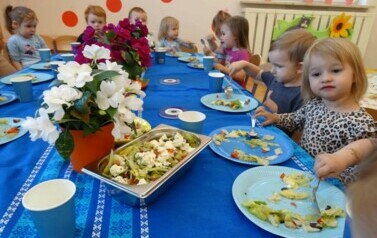 Grupka dzieci siedzi przy stoliku, w ręce trzymają łyżki i dotykają talerzy z warzywami.
