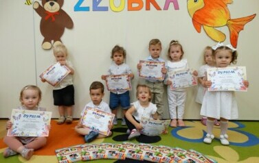 Grupa dzieci siedzi na dywanie. W ręce trzyma dyplomy. Nad nimi widnieje kolorowy napis "Pożegnanie żłobka".
