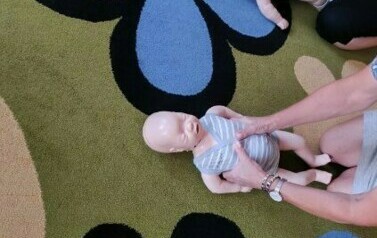 Na dywanie leżą fantomy szkoleniowe. Osoby dorosłe dotykają rękami ich klatki piersiowej.