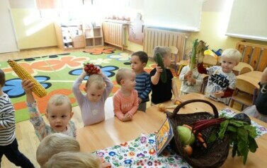 Grupka dzieci siedzi przy stoliku, na środku kt&oacute;rego leży książeczka i kosz z warzywami. Każde dziecko trzyma w ręce warzywko.