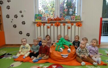 Na pomarańczowym kocu siedzi grupka dzieci. Obok nich stoi ogromna pomarańczowa dynia z zieloną czapką na głowie.