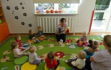 Sala zabaw-na dywanie siedzi osoba dorosła z grupką dzieci. W ręce trzyma pacynkę lalki i wilka.