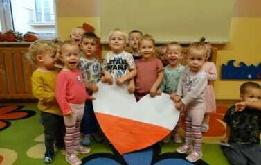 Grupka dzieci stoi na dywanie i w ręce trzyma ogromne serce w narodowych barwach.