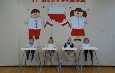 Grupka dzieci odświętnie ubrana siedzi w fotelikach do karmienia. W tle jasna ściana a na niej chłopiec i dziewczynka z biało-czerwonymi chorągiewkami i napis 11 LISTOPADA. 