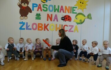Na krzesełkach siedzą dzieci. Przed nimi osoba dorosła z dużym oł&oacute;wkiem w ręce.W tle kolorowe obrazki i napis PASOWANIE NA ŻŁOBKOWICZA