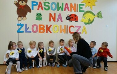 Na krzesełkach siedzą dzieci. Przed nimi osoba dorosła z dużym oł&oacute;wkiem w ręce.W tle kolorowe obrazki i napis PASOWANIE NA ŻŁOBKOWICZA