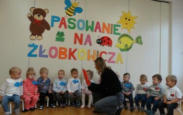 Na krzesełkach siedzą dzieci. Przed nimi osoba dorosła z dużym oł&oacute;wkiem w ręce.W tle kolorowe obrazki i napis PASOWANIE NA ŻŁOBKOWICZA