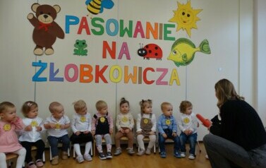 Na krzesełkach siedzą dzieci. Przed nimi osoba dorosła z dużym oł&oacute;wkiem w ręce.W tle kolorowe obrazki i napis PASOWANIE NA ŻŁOBKOWICZA.
