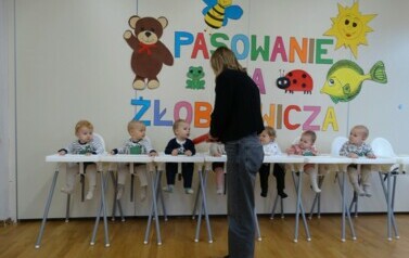 W białych fotelikach do karmienia siedzą dzieci. Tuż przy nich stoi osoba dorosła z dużym oł&oacute;wkiem w ręce. W tle kolorowe obrazki i napis PASOWANIE NA ŻŁOBKOWICZA. 