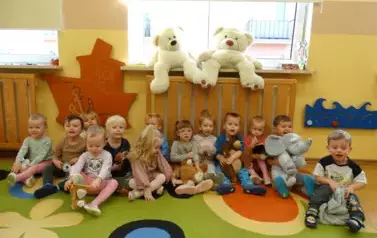 Na kolorowym dywanie siedzą dzieci. W ręce trzymają maskotki misie.