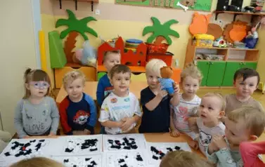 Przy stoliku stoją dzieci. Przed nimi leżą kartki z łaciatymi misiami.