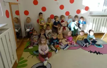 Na tle jasnej ściany w czerwone kropki stoją dzieci. W ręce trzymają maskotki misie.