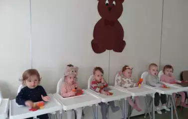 W białych fotelikach do karmienia siedzą dzieci. W tle obrazek z dużym brązowym misiem.