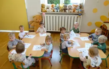 Przy stolikach siedzą dzieci. Przed nimi leżą białe kartkii z misiem.