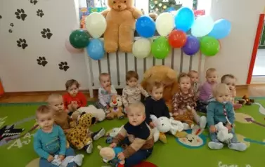Na kolorowym dywanie siedzą dzieci. Wręce trzymają pluszaki misie. W tle kolorowe balony z ogromnym misiem pluszakiem..