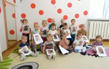 Na tle jasnej ściany w czerwone kropki stoją dzieci. W ręce trzymają obrazki z misiem.