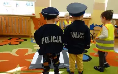 Dwaj chłpocy odwr&oacute;ceni tyłem stoją na dywanie. Na głowach mają czapki a na plecach napis POLICJA.