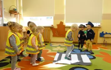 Na dywanie leży karton w biało-szare pasy, na kt&oacute;rym stoi dziecko. Obok stoją dwaj chłopcyw przebraniu policjanta. Jeden z nich trzyma w ręku znak z zielonym k&oacute;łeczkiem..