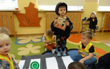 Dzieci w odblaskowych kamizelkach siedzą na dywanie. Przed nimi leży karton w biało-szare pasy. Obok stoi chłopiec w czapce z daszkiem i w ręce trzyma książeczkę.
