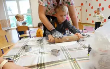 Dziecko z osobą dorosłą stoją przy stoliku, na kt&oacute;rym leży okrągły placek. W ręce trzymają wałek do wałkowania ciasta.