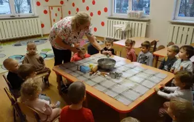 Przy stoliku siedzą dzieci. Obok nich stoi osoba dorosła. Nad miską trzyma przechylony słoiczek.