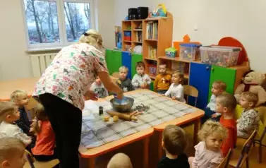Przy stoliku siedzą dzieci. Obok nich stoi osoba dorosła. Nad miską trzyma niebieską torebkę.