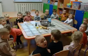 Przy stoliku siedzą dzieci a na nim leży wałek do ciasta, miska, foremki.