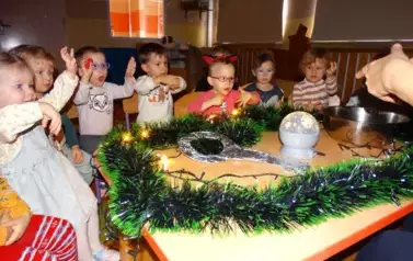 Przy stoliku siedzą dzieci z uniesionymi rączkami a na nim stoi miska, świecąca kula  i duży klucz. 