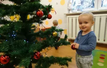 Dziecko stoi przy choince, w ręce trzyma czerwoną bombkę.