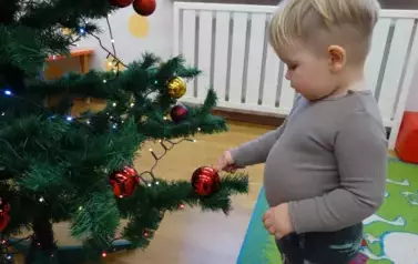 Dziecko stoi przy choince, rączką dotyka bombki zawieszonej na choince.