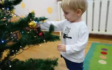 Dziecko stoi przy choince, rączką dotyka bombki zawieszonej na choince.