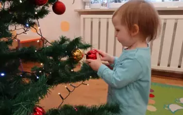 Dziecko stoi przy choince, w ręce trzyma czerwoną bombkę i dotyka zielonej gałązki.