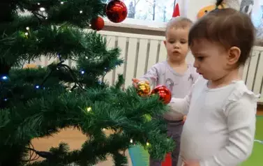 Dwoje dzieci stoi przy choince, w ręce trzymają kolorowe bombki i dotykają zielonych gałązek.