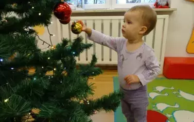Dziecko stoi przy choince, w ręce trzyma złotą bombkę i dotyka zielonej gałązki.