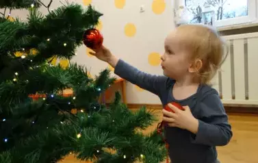 Dziecko stoi przy choince. W jednej rączce trzyma bombkę a drugą dotyka bombki zawieszonej na choince.
