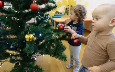 Dw&oacute;jka dzieci stoi przy choince, w ręce trzyma czerwone bombki.