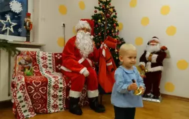 Na pierwszym planie stoi dziecko z dzwoneczkiem w ręce. W tle kolorowa choinka i siedzący Mikołaj z laską i workiem w ręce.
