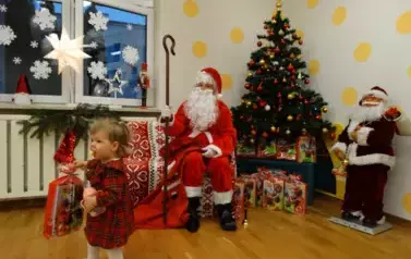 Dziewczynka w kolorowej sukience trzyma w ręce paczkę. W tle siedzący Mikołaj z laską w ręce i zielona choinka z kolorowymi światełkami..