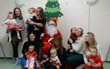 Mikołaj siedzi na krześle z laską w ręce. Tuż obok siedzi grupka dorosłych z dziećmi na rękach. W tle zielona choinka i gwiazdki. 