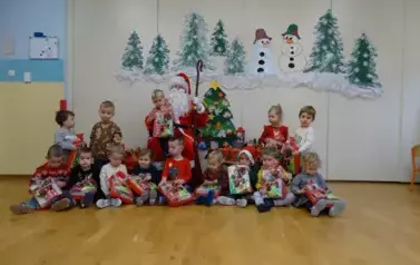 Mikołaj siedzi na krześle z dzieckiem na kolanach. Tuż obok na podłodze siedzi grupka dzieci z kolorowymi paczkami. Wtle zielone choinki i białe bałwanki.