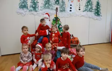Mikołaj z dzieckiem na kolanach, obok niego na podłodze siedzi grupka dzieci. W tle zielone choinki i białe mikołajki.