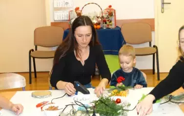 Przy stoliku siedzi chłopiec. W ręce trzyma czerwoną jarzębinę. Obok siedzi kobieta, w ręce trzyma pistolet z klejem.