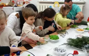 Grupa dorosłych os&oacute;b z dziećmi siedzi przy stoliku. Na nim leżą zielone gałązki świerku, srebrne bombki i plasterki cytryny.